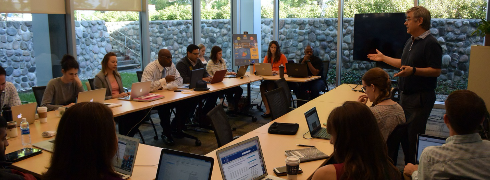 approximately 15 people seated at a U-shaped table listening to a standing presenter