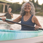 Smiling woman with glasses and grey hair paddles a kayak on a river in the foreground with a man in kayak paddling alongside her. There is a bridge in the distant background.