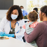 A concerned young mother talks with a female pediatrician during an appointment at a free clinic. The pediatrician is taking notes as the young mom talks about her daughter's symptoms.