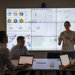 A group of three researchers with laptops sit around a conference table studying data on a projected screen. A fourth researcher stands in front of the screen and addresses the three at the table.