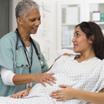 Black female doctor examining pregnant patient's belly in exam room.